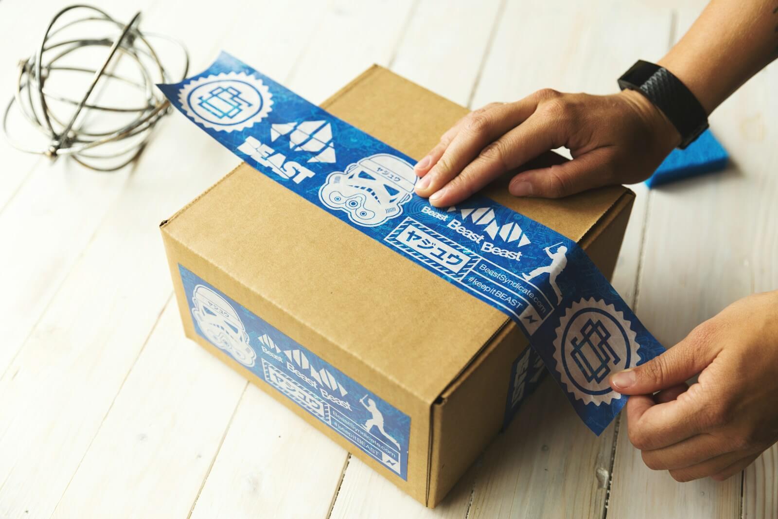 Hands sealing a cardboard box with blue tape — preparing a courier parcel for dispatch